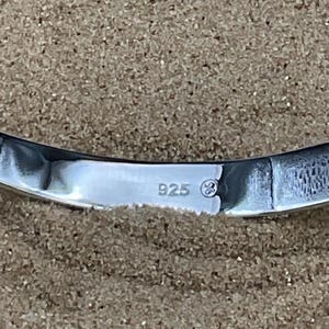 May include: Close-up of a curved, polished silver object. The object has a reflective surface and the number "925" is stamped on it, indicating its silver content. The background is a textured, neutral color.
