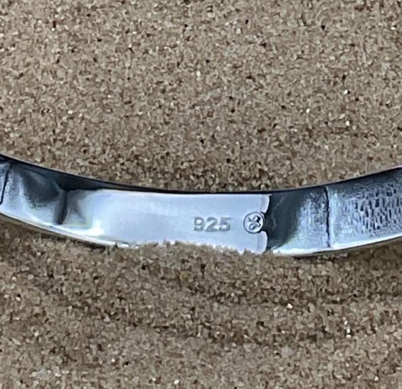 May include: Close-up of a curved, polished silver object. The object has a reflective surface and the number "925" is stamped on it, indicating its silver content. The background is a textured, neutral color.