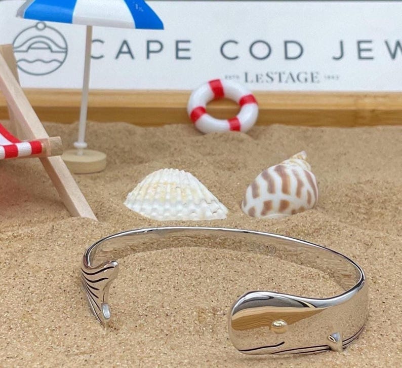 May include: A silver whale tail cuff bracelet with a gold accent, resting on sand with seashells and miniature beach accessories. The bracelet is a nautical-themed jewelry piece.