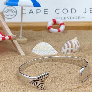 May include: A silver cuff bracelet with a fish tail design. The bracelet is open, with a tail fin on one end and a rounded end on the other. The bracelet is on a sandy surface with seashells and a miniature beach scene in the background.