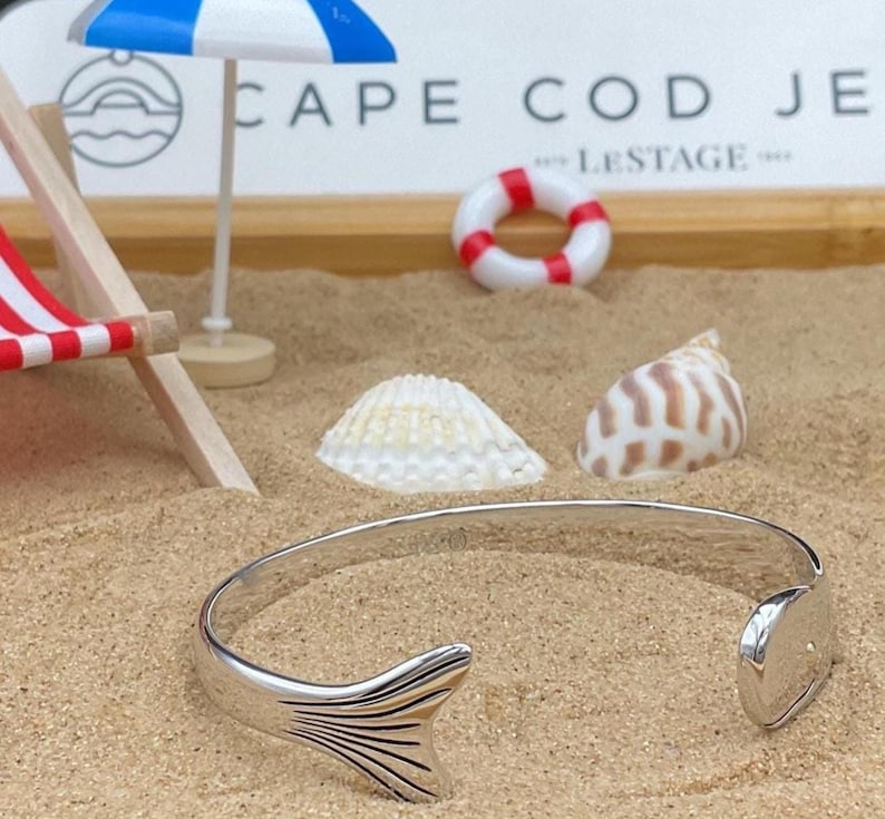May include: A silver cuff bracelet with a fish tail design. The bracelet is open, with a tail fin on one end and a rounded end on the other. The bracelet is on a sandy surface with seashells and a miniature beach scene in the background.