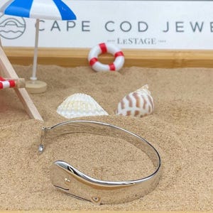 May include: A silver whale tail bangle bracelet with a gold accent, displayed on sand with a miniature beach scene. The scene includes a miniature beach chair, a life preserver, seashells, and a small beach umbrella. The text "CAPE COD JEWEL" is visible.