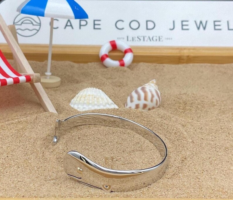 May include: A silver whale tail bangle bracelet with a gold accent, displayed on sand with a miniature beach scene. The scene includes a miniature beach chair, a life preserver, seashells, and a small beach umbrella. The text "CAPE COD JEWEL" is visible.
