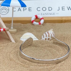 May include: A silver cuff bracelet rests on sand, with a miniature beach scene in the background. The scene includes a small beach umbrella, a life preserver, and seashells. The text "CAPE COD JEW" is visible.