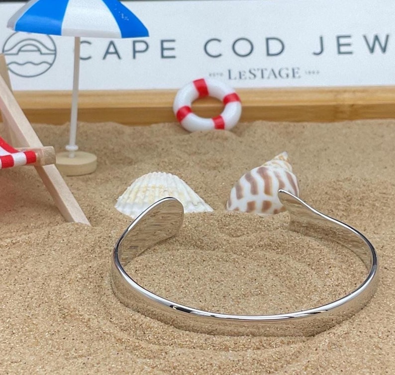 May include: A silver cuff bracelet rests on sand, with a miniature beach scene in the background. The scene includes a small beach umbrella, a life preserver, and seashells. The text "CAPE COD JEW" is visible.