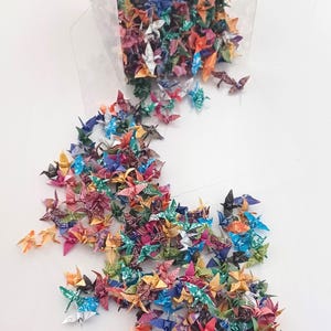May include: A clear plastic container filled with colorful origami paper cranes. The cranes are spilling out of the container onto a white surface. The text "Pattern A" is visible at the bottom of the image.