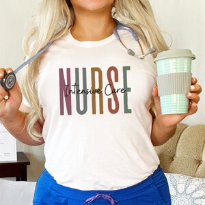 May include: A white t-shirt with the text "NURSE Intensive Care" in colorful letters. The person wearing the shirt is holding a stethoscope and a reusable coffee cup.