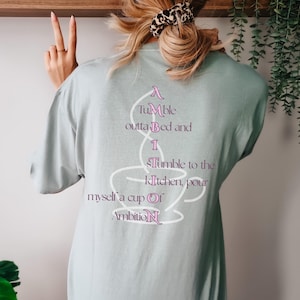 Op de afbeelding: Een licht saliegroen t-shirt met een witte omtrek van een koffiekopje op de achterkant. De tekst luidt: A Tumble outta Bed and I Tumble to the kitchen, pour myself a cup Of AmbitioN, waarbij de letters A, I, S, O en N roze zijn gemarkeerd.