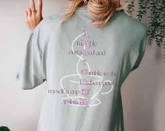 May include: A light sage green t-shirt featuring a white outline of a coffee cup on the back. The text reads: A Tumble outta Bed and I Tumble to the kitchen, pour myself a cup Of AmbitioN, with the letters A, I, S, O, and N highlighted in pink.
