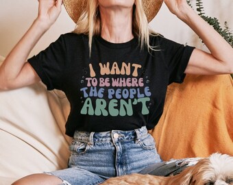 Op de afbeelding: Een vrouw draagt een zwart T-shirt met de tekst "I WANT TO BE WHERE THE PEOPLE AREN'T" in regenboogkleuren en zit op een witte bank met een hond. Ze draagt een strohoed en een blauwe spijkerbroek.