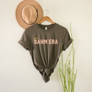Puede incluir: Una camiseta verde oscuro con el texto "IN MY SAHM ERA" en letras rosas y amarillas.