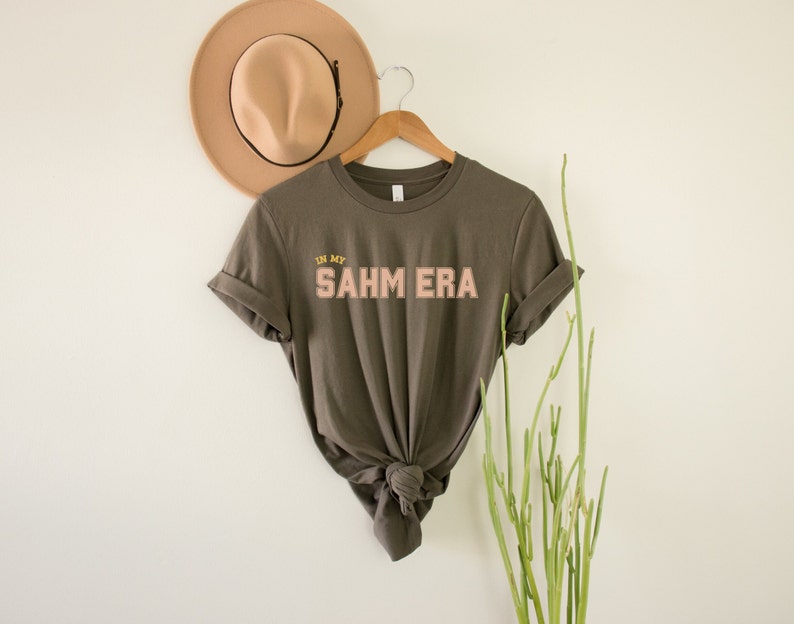 Puede incluir: Una camiseta verde oscuro con el texto "IN MY SAHM ERA" en letras rosas y amarillas.