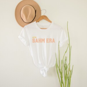 Puede incluir: Camiseta blanca con el texto "In my SAHM ERA" en letras color melocot&oacute;n y amarillo.