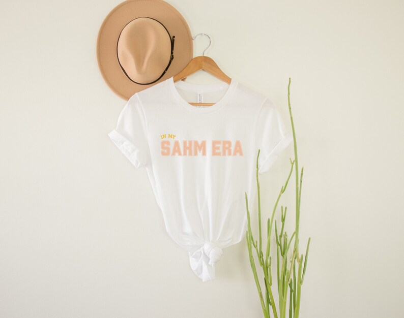 Puede incluir: Camiseta blanca con el texto "In my SAHM ERA" en letras color melocot&oacute;n y amarillo.