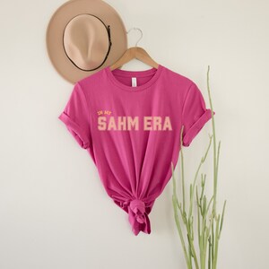 Puede incluir: Una camiseta rosa con el texto "In My SAHM ERA" en letras naranja y amarillas.