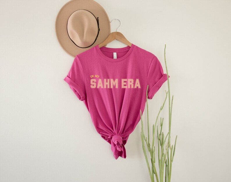 Puede incluir: Una camiseta rosa con el texto "In My SAHM ERA" en letras naranja y amarillas.