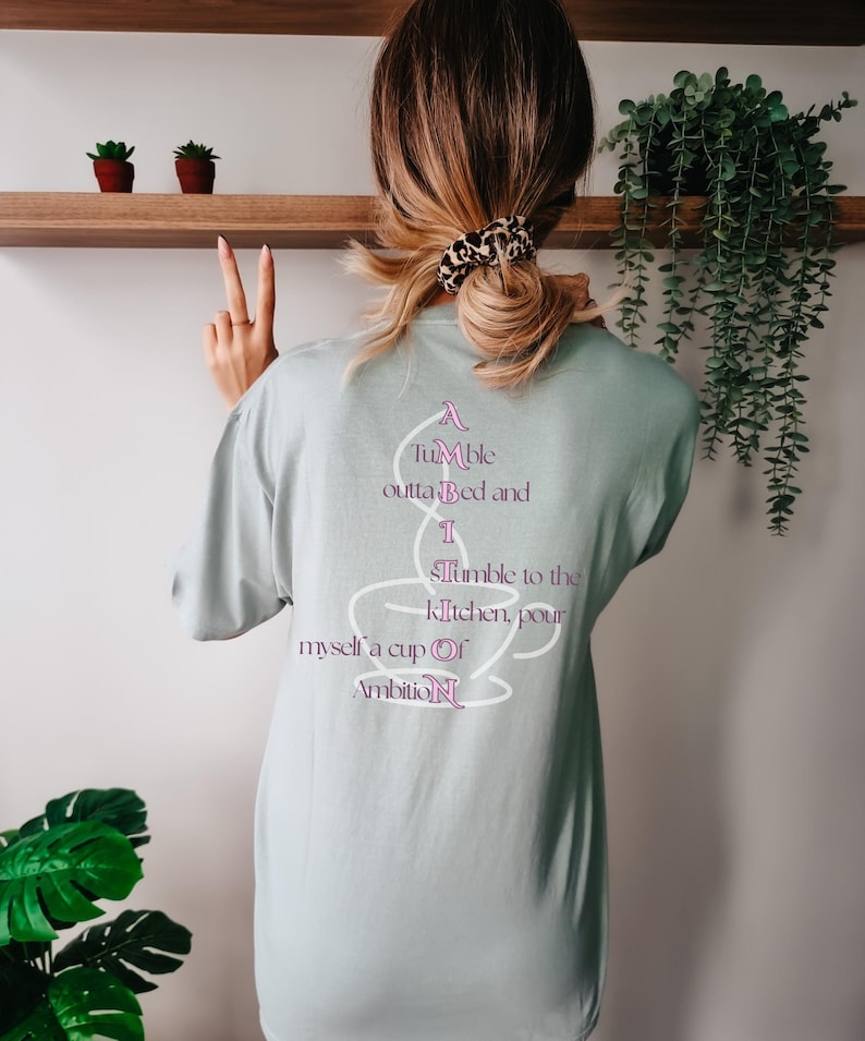 Op de afbeelding: Een licht saliegroen t-shirt met een witte omtrek van een koffiekopje op de achterkant. De tekst luidt: A Tumble outta Bed and I Tumble to the kitchen, pour myself a cup Of AmbitioN, waarbij de letters A, I, S, O en N roze zijn gemarkeerd.