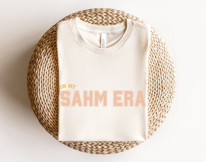 Puede incluir: Una camiseta blanca con el texto "In My SAHM Era" impreso en melocot&oacute;n y amarillo.