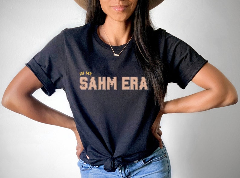 Puede incluir: Una camiseta negra con el texto "In My SAHM ERA" en letras doradas y marrones.