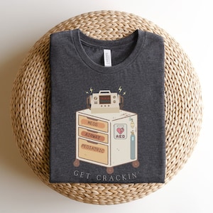 May include: Dark heather grey t-shirt with a cartoon illustration of a medical cart with the text "MEDS, AIRWAY, PEDIATRIC, AED, GET CRACKIN'" printed on it.