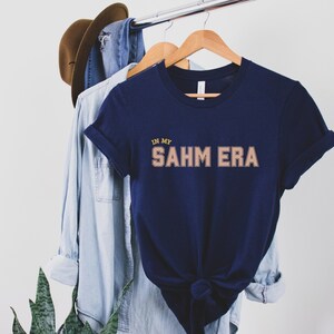Puede incluir: Una camiseta azul marino con el texto "IN MY SAHM ERA" en color marr&oacute;n claro con contorno amarillo. La camiseta est&aacute; colgada de una percha de madera.
