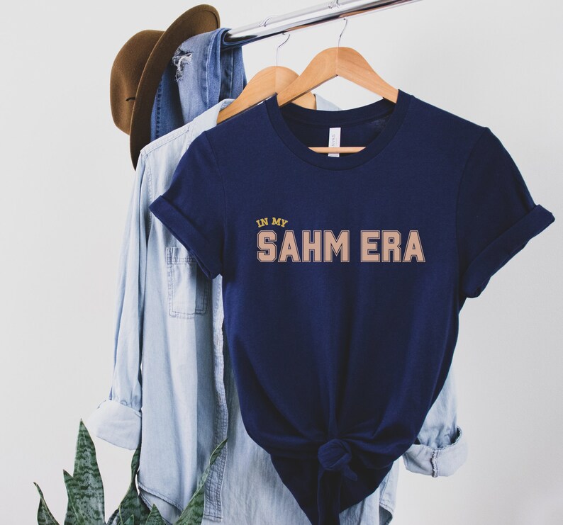 Puede incluir: Una camiseta azul marino con el texto "IN MY SAHM ERA" en color marr&oacute;n claro con contorno amarillo. La camiseta est&aacute; colgada de una percha de madera.