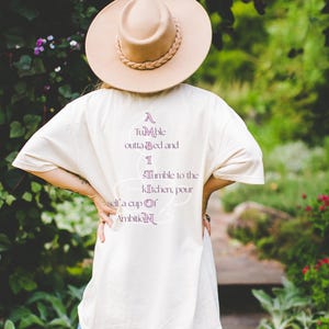 Op de afbeelding: Een beige t-shirt met de paarse tekst A Tumble outta Bed and I Tumble to the kitchen, pour self a cup of Ambition, gecombineerd met een beige hoed met brede rand en gevlochten band en een blauwe spijkerbroek, gezien vanaf de achterkant.