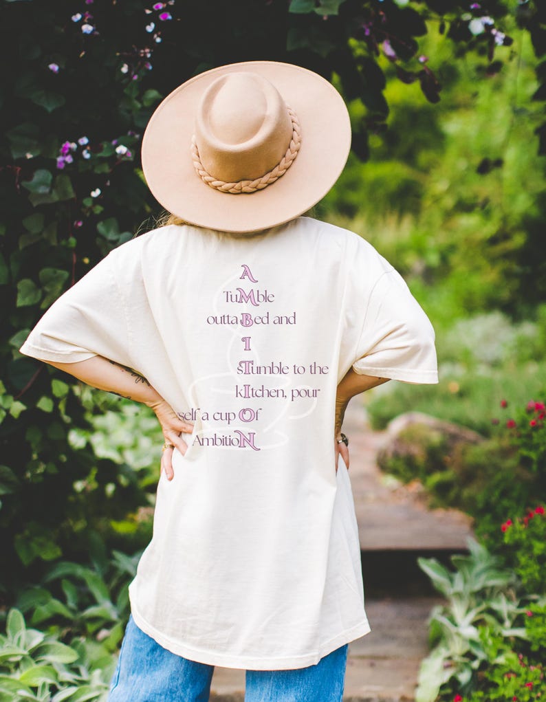 Op de afbeelding: Een beige t-shirt met de paarse tekst A Tumble outta Bed and I Tumble to the kitchen, pour self a cup of Ambition, gecombineerd met een beige hoed met brede rand en gevlochten band en een blauwe spijkerbroek, gezien vanaf de achterkant.
