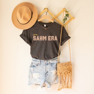 Puede incluir: Una camiseta gris oscuro con el texto "In My SAHM Era" en letras rosas y doradas. La camiseta est&aacute; colgada en una percha de madera con un sombrero de paja y un bolso tejido.
