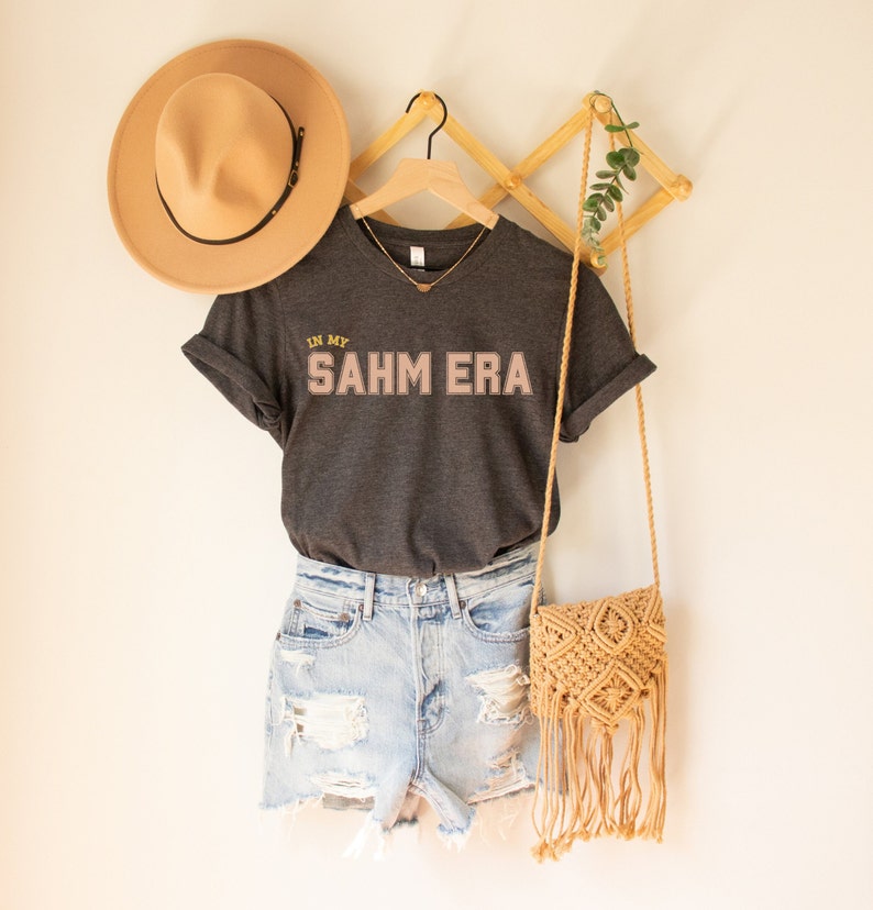 Puede incluir: Una camiseta gris oscuro con el texto "In My SAHM Era" en letras rosas y doradas. La camiseta est&aacute; colgada en una percha de madera con un sombrero de paja y un bolso tejido.