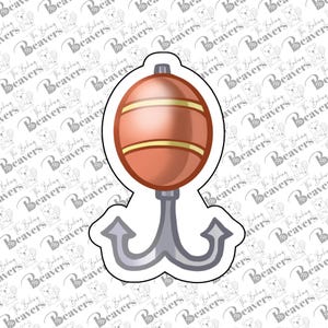 May include: A white cookie cutter shaped like a fishing float with a rounded orange body featuring two horizontal yellow stripes, attached to a gray double-pronged anchor hook at the bottom, outlined with a thick white border.