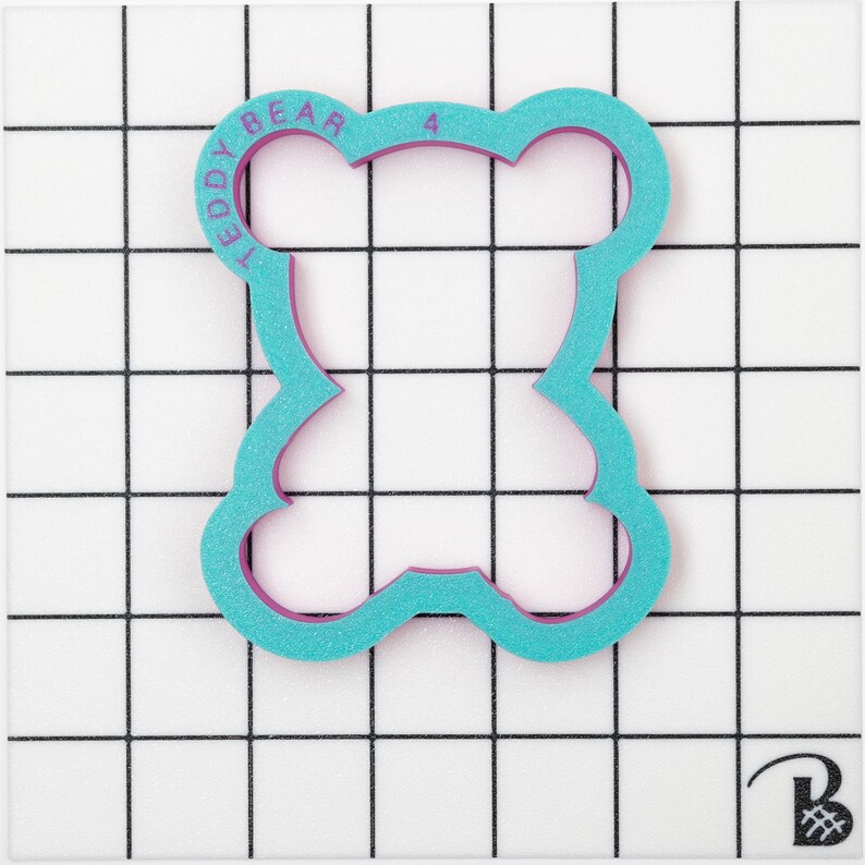 May include: A turquoise blue teddy bear cookie cutter with a pink outline. The cutter is on a white grid background. The text "TEDDY BEAR" is printed on the cutter.