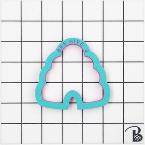 May include: A teal and purple bee hive shaped cookie cutter. The text "BEE HIVE 3" is printed on the top of the cutter. The cutter is on a white surface with a black grid pattern.