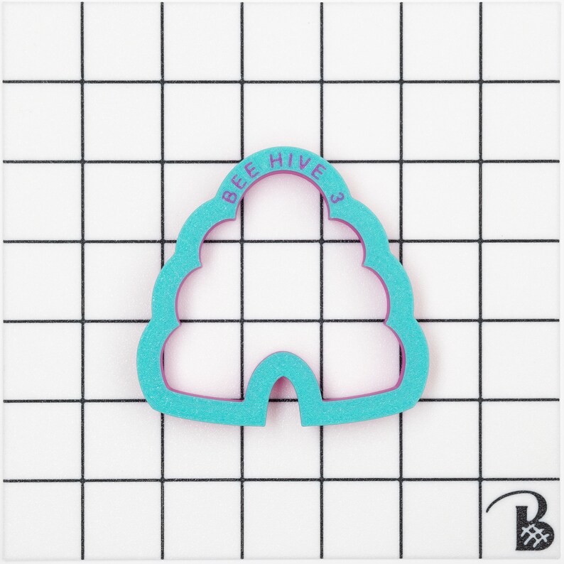 May include: A teal and purple bee hive shaped cookie cutter. The text "BEE HIVE 3" is printed on the top of the cutter. The cutter is on a white surface with a black grid pattern.