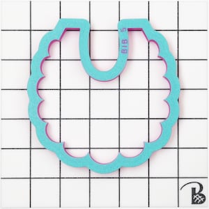 May include: A teal and pink bib-shaped cookie cutter with a scalloped edge. The cutter has the text "BIB 5" printed on it. The cutter is on a white grid background.