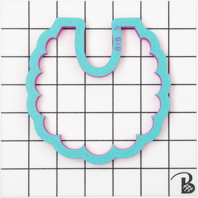 May include: A teal and pink bib-shaped cookie cutter with a scalloped edge. The cutter has the text "BIB 5" printed on it. The cutter is on a white grid background.