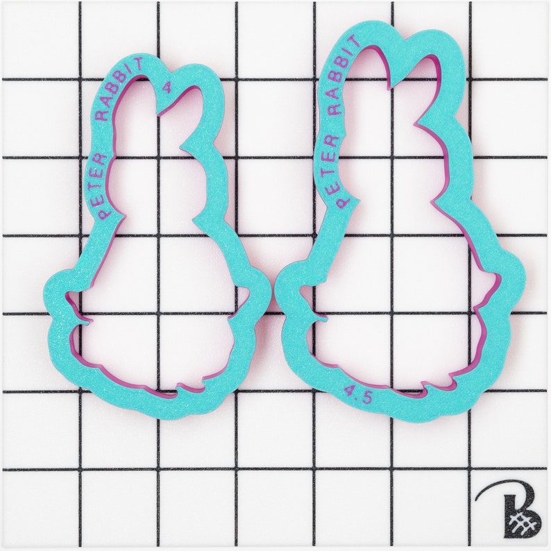 May include: Two Peter Rabbit cookie cutters. The cutters are teal with a pink interior and feature the words "PETER RABBIT" in pink. One cutter is labeled "4" and the other is labeled "4.5". The cutters are on a white grid background.