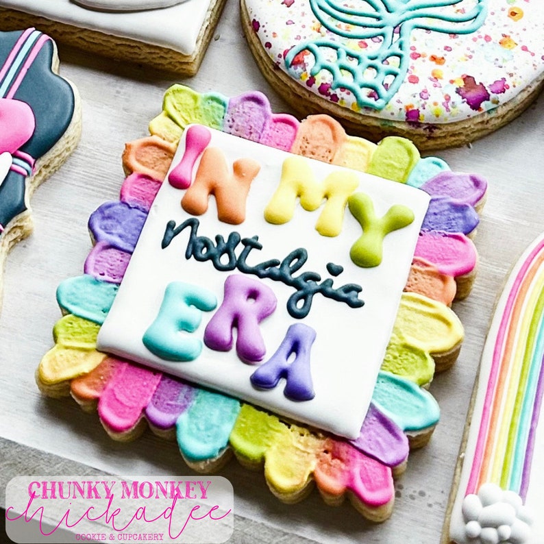 May include: A square-shaped cookie decorated with colourful icing. The cookie has a white background with the text "In My Nostalgia Era" written in black icing. The border of the cookie is decorated with a rainbow of colours.