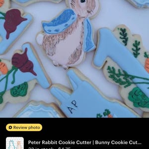 May include: Decorated sugar cookies with a Peter Rabbit theme. The cookies are in the shape of a bunny, carrots, and other shapes, with blue and white icing. The bunny cookie has a blue scarf. The cookies are decorated with intricate details.