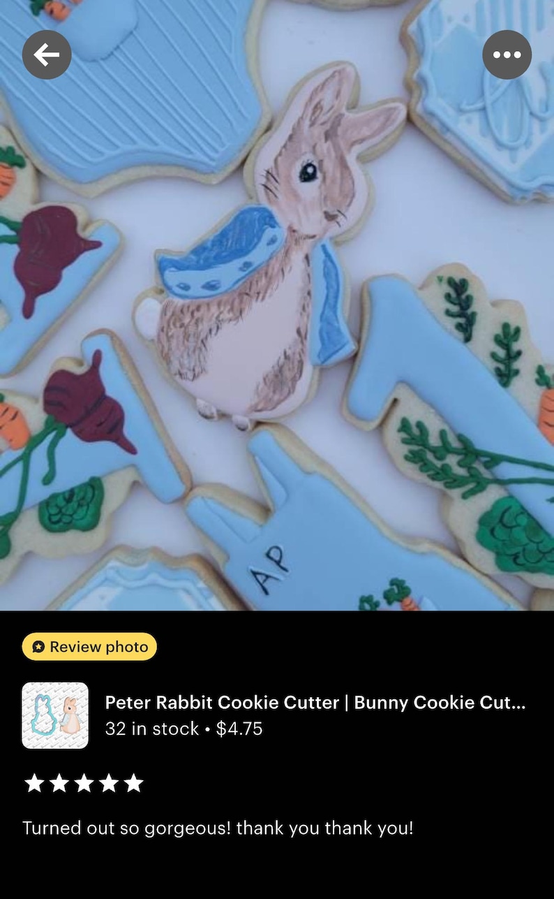 May include: Decorated sugar cookies with a Peter Rabbit theme. The cookies are in the shape of a bunny, carrots, and other shapes, with blue and white icing. The bunny cookie has a blue scarf. The cookies are decorated with intricate details.