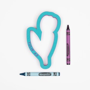 Op de afbeelding: Een turquoise blauwe plastic koekjesvormer in de vorm van een tulp met de tekst "TULIP 4.5" aan de zijkant. Twee kleurpotloden, &eacute;&eacute;n paars en &eacute;&eacute;n blauw, zijn ook afgebeeld.