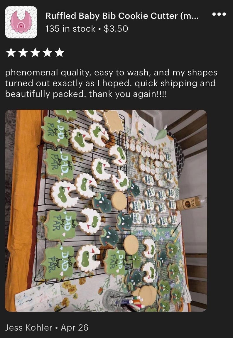 May include: A display of decorated sugar cookies, featuring golf course and baby bib designs. The cookies are iced in green, white, and beige, and arranged on wire racks. Some cookies have the text "Best Dad by Par".