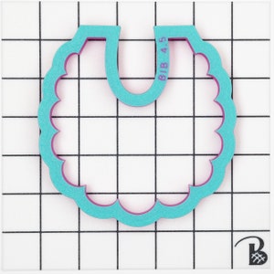 May include: A teal bib-shaped cookie cutter with a scalloped edge and a purple interior. The text "BIB 4.5" is printed on the top right. The cutter is on a white surface with a black grid pattern.