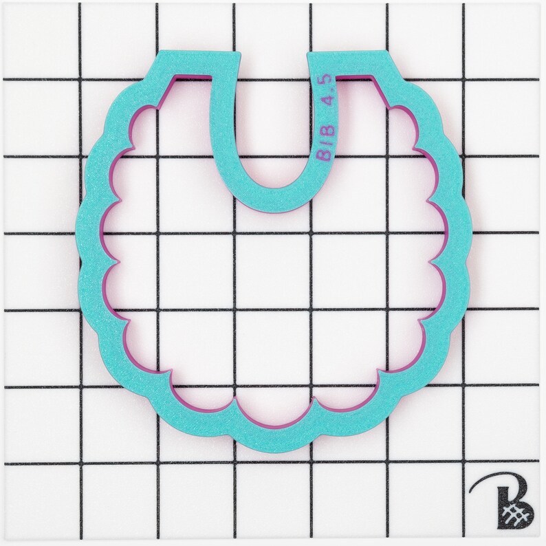 May include: A teal bib-shaped cookie cutter with a scalloped edge and a purple interior. The text "BIB 4.5" is printed on the top right. The cutter is on a white surface with a black grid pattern.