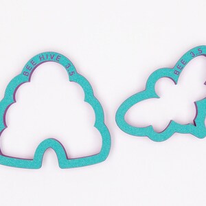May include: Two teal cookie cutters, one shaped like a beehive, the other like a bee. The beehive cutter has the text "BEE HIVE 3.5" and the bee cutter has the text "BEE 3.5". The cutters are for baking.