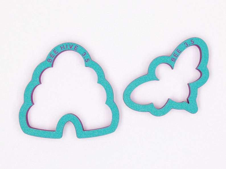 May include: Two teal cookie cutters, one shaped like a beehive, the other like a bee. The beehive cutter has the text "BEE HIVE 3.5" and the bee cutter has the text "BEE 3.5". The cutters are for baking.