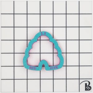 May include: A teal beehive-shaped cookie cutter with the words "BEE HIVE" and the number "2.5" in purple. The cutter is on a white surface with a black grid. The measurement is 6.35 cm.