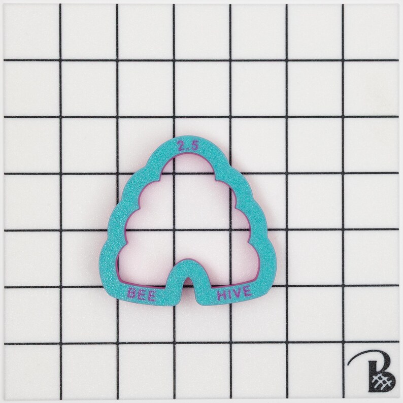 May include: A teal beehive-shaped cookie cutter with the words "BEE HIVE" and the number "2.5" in purple. The cutter is on a white surface with a black grid. The measurement is 6.35 cm.