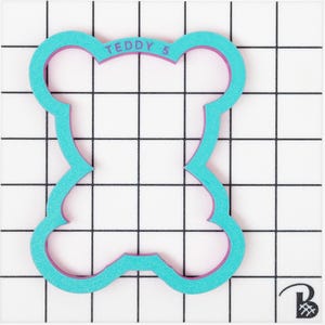May include: A turquoise blue cookie cutter shaped like a teddy bear with a pink outline. The cookie cutter has the text "TEDDY 5" on the top.