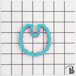 May include: A teal and purple cookie cutter shaped like a bib, with a scalloped edge. The text "BIB" and "2.5" are visible. The cutter is on a white surface with a black grid pattern.